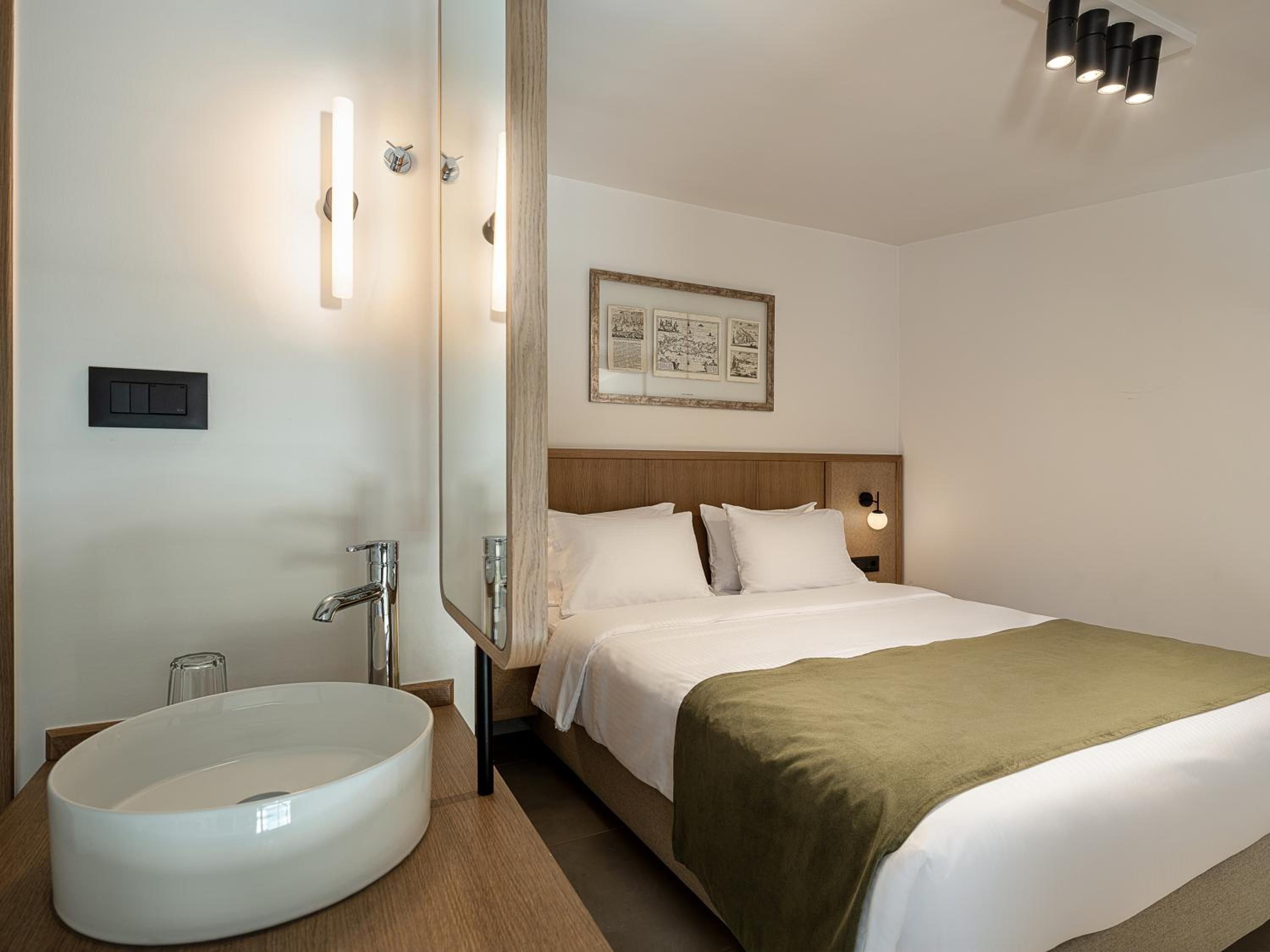 A hotel room with a large round sink, a bed with a green blanket, a wooden headboard, and a framed picture on the wall.