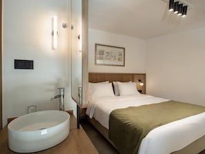 Une chambre d’hôtel avec un grand lavabo rond, un lit recouvert d’une couverture verte, une tête de lit en bois et un tableau encadré accroché au mur.