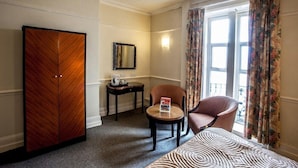 Una habitación de hotel con cama, armario, silla, mesita pequeña, mesita de noche, espejo y ventana con cortinas.