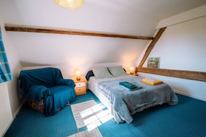 A bedroom with a sloped ceiling, a bed with a blue blanket, a chair, bedside tables with lamps, and a painting on the wall.