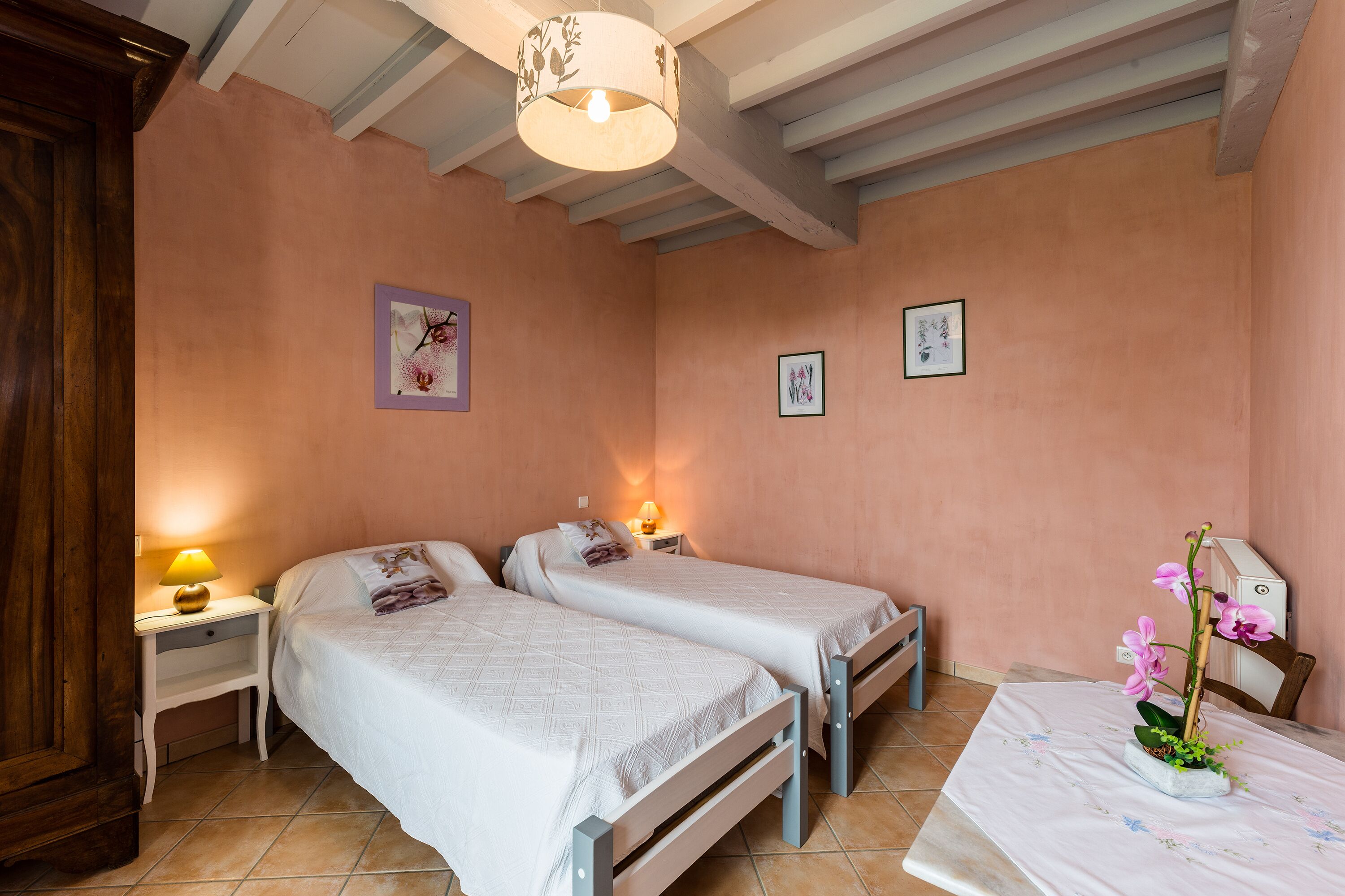 A bedroom with two beds, a nightstand with a lamp, and a small table with a flower arrangement.