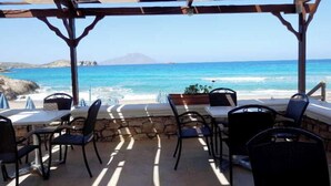 An outdoor seating area with tables and chairs, overlooking a beach and ocean. - Royal Beach Hotel (Karpathos)