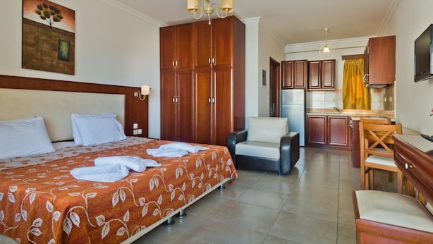 A hotel room with a bed, a desk, a chair, a kitchenette, and a painting on the wall. - Royal Beach Hotel (Karpathos)