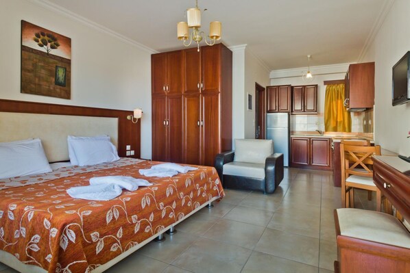 A hotel room with a bed, a desk, a chair, a kitchenette, and a painting on the wall.