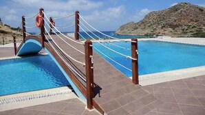 A pool area with a rope bridge, a lifebuoy, and a view of the sea and hills. - Royal Beach Hotel (Karpathos)