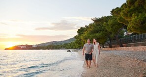 A couple walking along a pebble beach with a sunset in the background. - Heritage Hotel Kastelet (Tucepi)