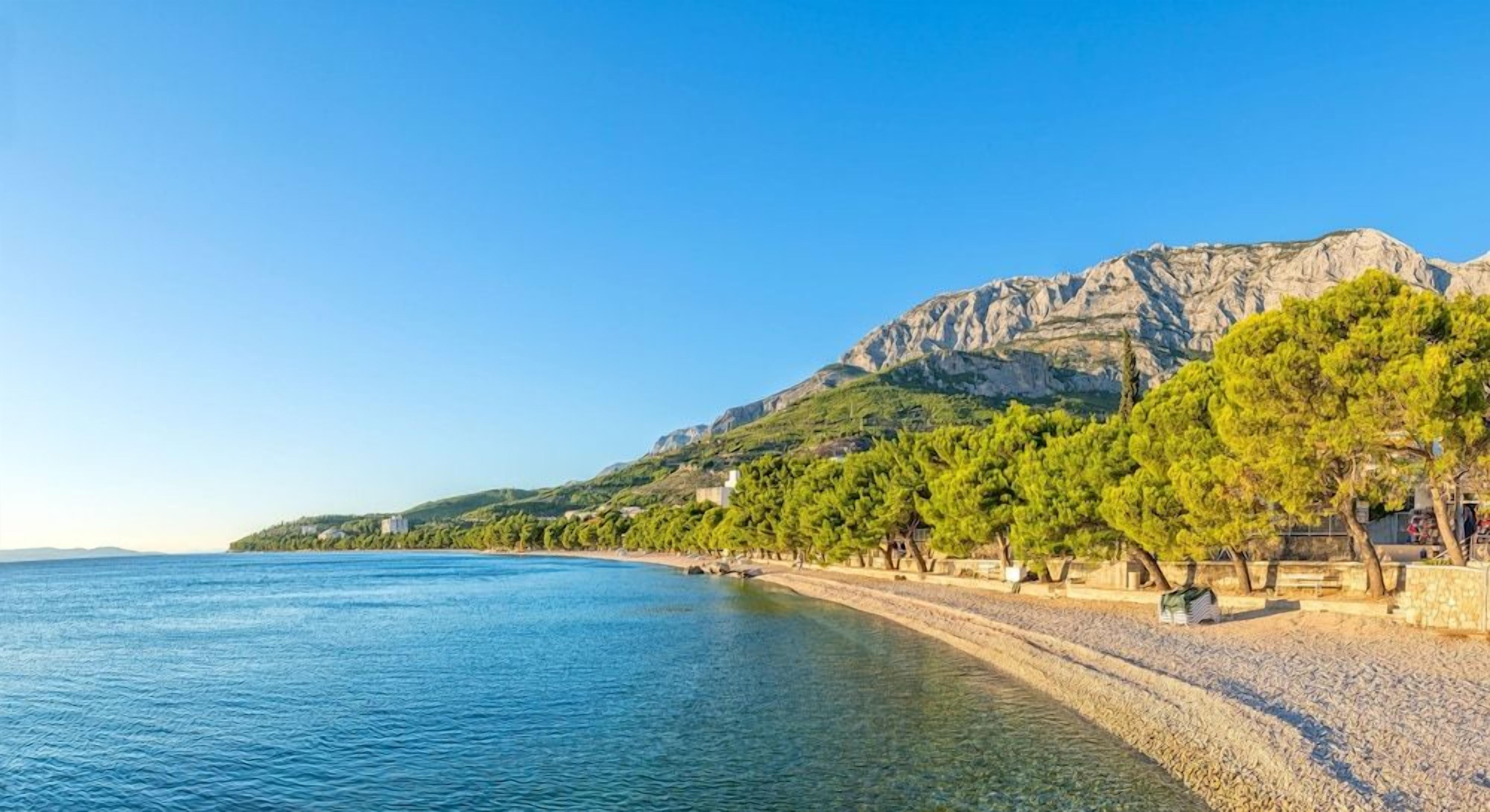 Un paisaje costero con mar azul cristalino, una playa de guijarros y una montaña al fondo.