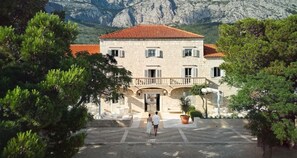 A building with a red-tiled roof, a balcony, and a paved courtyard with trees. - Heritage Hotel Kastelet (Tucepi)