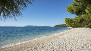 A pebble beach with clear water, a distant shoreline, and a tree-lined path. - Heritage Hotel Kastelet (Tucepi)