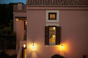 A building with a pink facade, a window with wooden shutters, and wall-mounted lights. - Eliathos Residence Houses Archanes (Archanes-Asterousia)