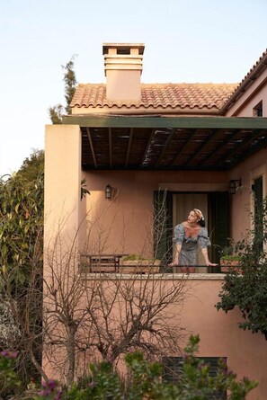 Une personne se tient sur un balcon, d’où l’on peut admirer un bâtiment et la végétation environnante.