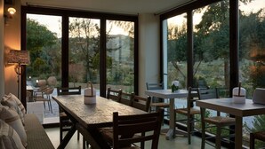 Uma área de refeições com uma mesa grande e cadeiras, janelas amplas com vista para árvores e colinas, e um sofá com almofadas.