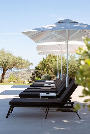 A row of sun loungers under a large umbrella, with a pool and trees in the background. - Eliathos Residence Houses Archanes (Archanes-Asterousia)