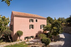 Un bâtiment à deux étages, doté d’un toit recouvert de tuiles, de nombreuses fenêtres avec volets et d’une cour pavée de gravier.