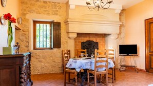 A dining area with a brick fireplace, wooden chairs, a table with a white tablecloth, a chandelier, and a television on a stand.