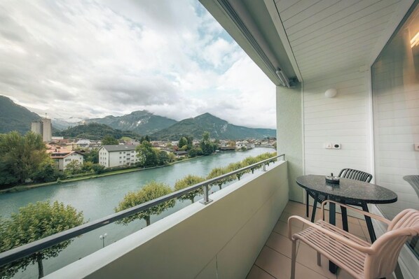 Un balcón con vistas a un río y montañas, una mesa con sillas y una barandilla.