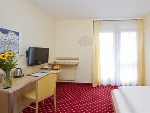 A hotel room with a bed, a desk with a chair, a television, a vase with flowers, and a window with curtains. - Aparthotel Goldey (Unterseen)