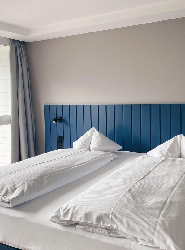 A hotel room with a bed, a headboard, a window with curtains, and a wall-mounted light fixture. - Aparthotel Goldey (Unterseen)