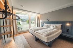 A bedroom with a bed, a window with a view of buildings and mountains, a wooden ladder, and a wall-mounted light fixture. - Aparthotel Goldey (Unterseen)