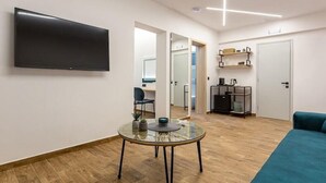 A modern living room with a flat-screen TV, a glass table, a sofa, and a kitchen area in the background. - Sirocco Hotel (Corfu)