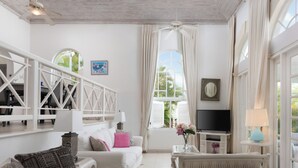 A spacious room with a white ceiling, a large window with curtains, a flat-screen TV, and a white sofa with cushions. - Royal Westmoreland Cassia Heights 29 - by Zenbreak (Westmoreland)