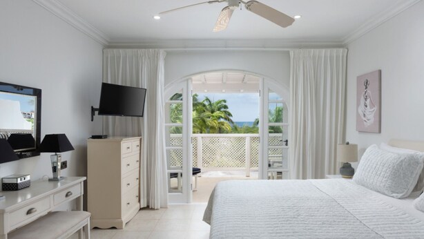 A bedroom with a bed, a TV, a dresser, and a balcony with a view of palm trees. - Royal Westmoreland Cassia Heights 29 - by Zenbreak (Westmoreland)