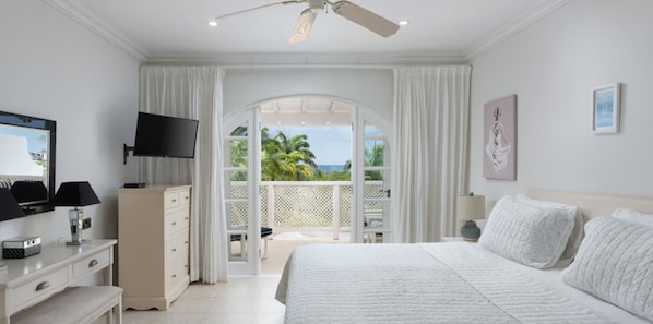 A bedroom with a bed, a TV, a dresser, and a balcony with a view of palm trees.