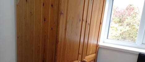 A wooden wardrobe in a room with a window, a bed with a floral bedspread, and a power outlet on the wall.