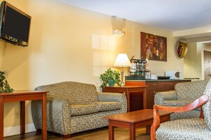 A living room with patterned sofas, a wooden coffee table, a flat-screen TV, a lamp, a painting, and a kitchen area with a microwave.