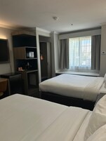 A hotel room with two beds, a desk, a minibar, and a window with curtains.