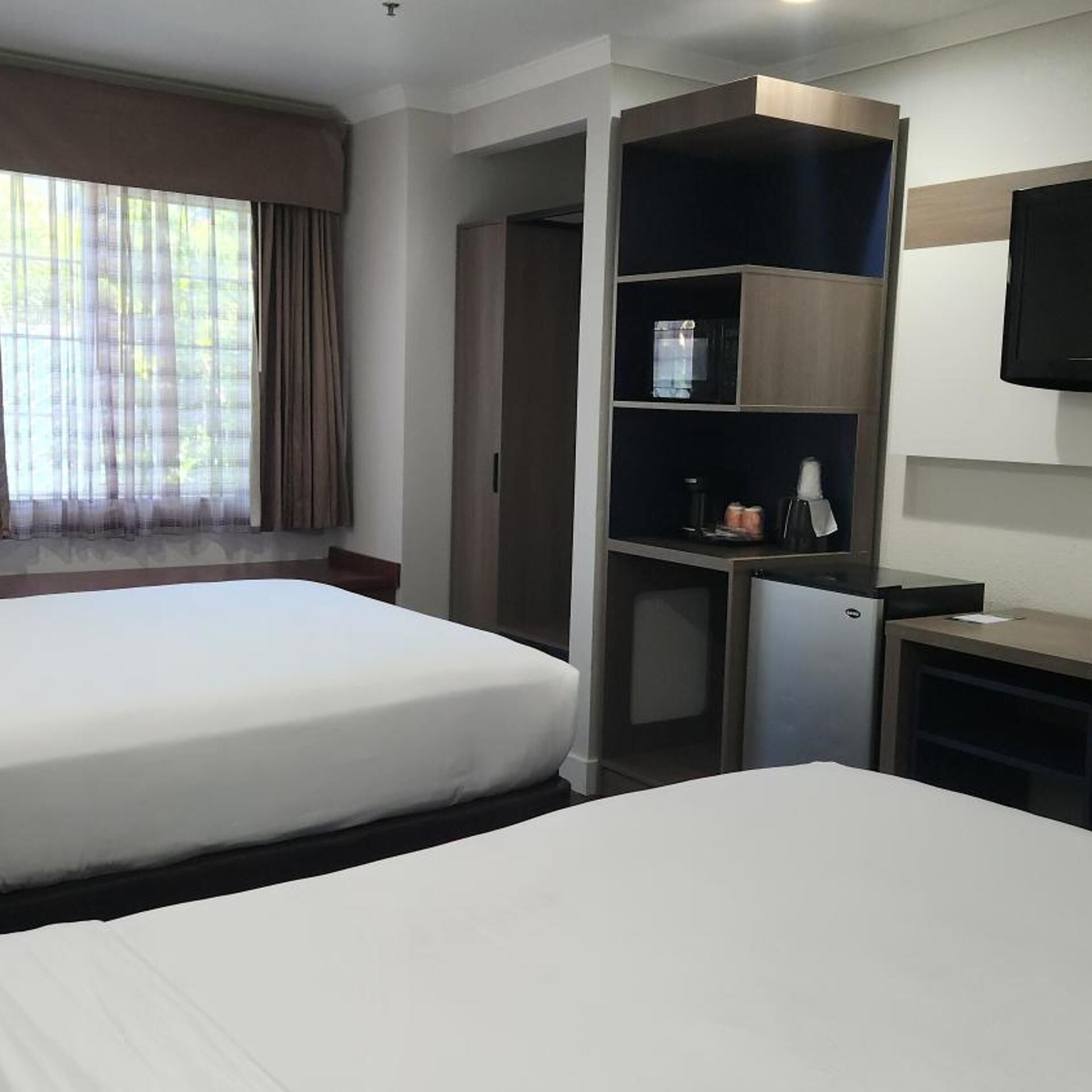 A hotel room with two beds, a desk with a computer, a TV mounted on the wall, and a window with curtains.
