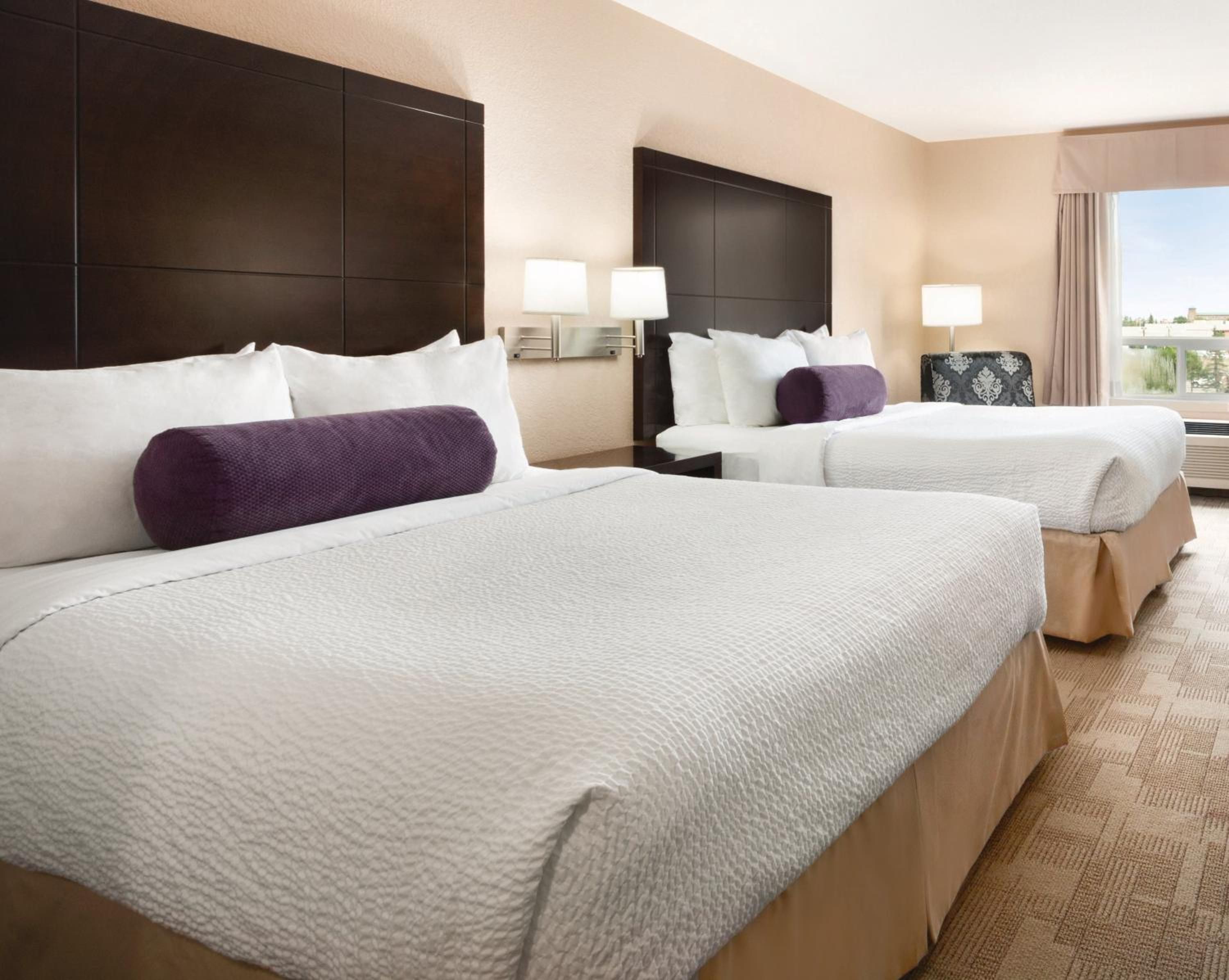 A hotel room with two beds, each with white bedding and a purple pillow. There are wall-mounted lamps and a window with curtains.