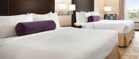 A hotel room with two beds, each with white bedding and a purple pillow. There are wall-mounted lamps and a window with curtains.