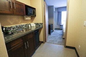 A compact kitchen with a microwave, sink, and dishwasher. A hallway leads to a bedroom with a window and curtains.
