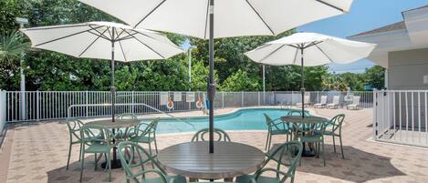 An outdoor patio with a pool, surrounded by a white fence, featuring green chairs and large umbrellas.