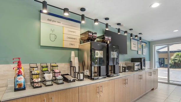 A modern coffee station with a coffee machine, creamer, sweetener, and a sign for adding your own flavor. - Sleep Inn & Suites Panama City Beach East (Panama City Beach)
