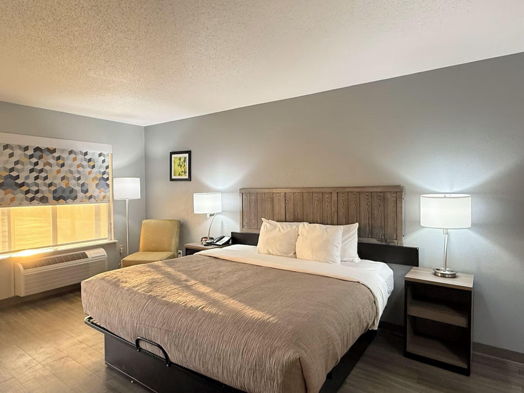 A hotel room with a bed, two bedside tables, a chair, a wall-mounted heater, and a decorative wall panel.