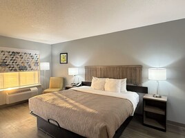 A hotel room with a bed, two bedside tables, a chair, a wall-mounted heater, and a decorative wall panel.