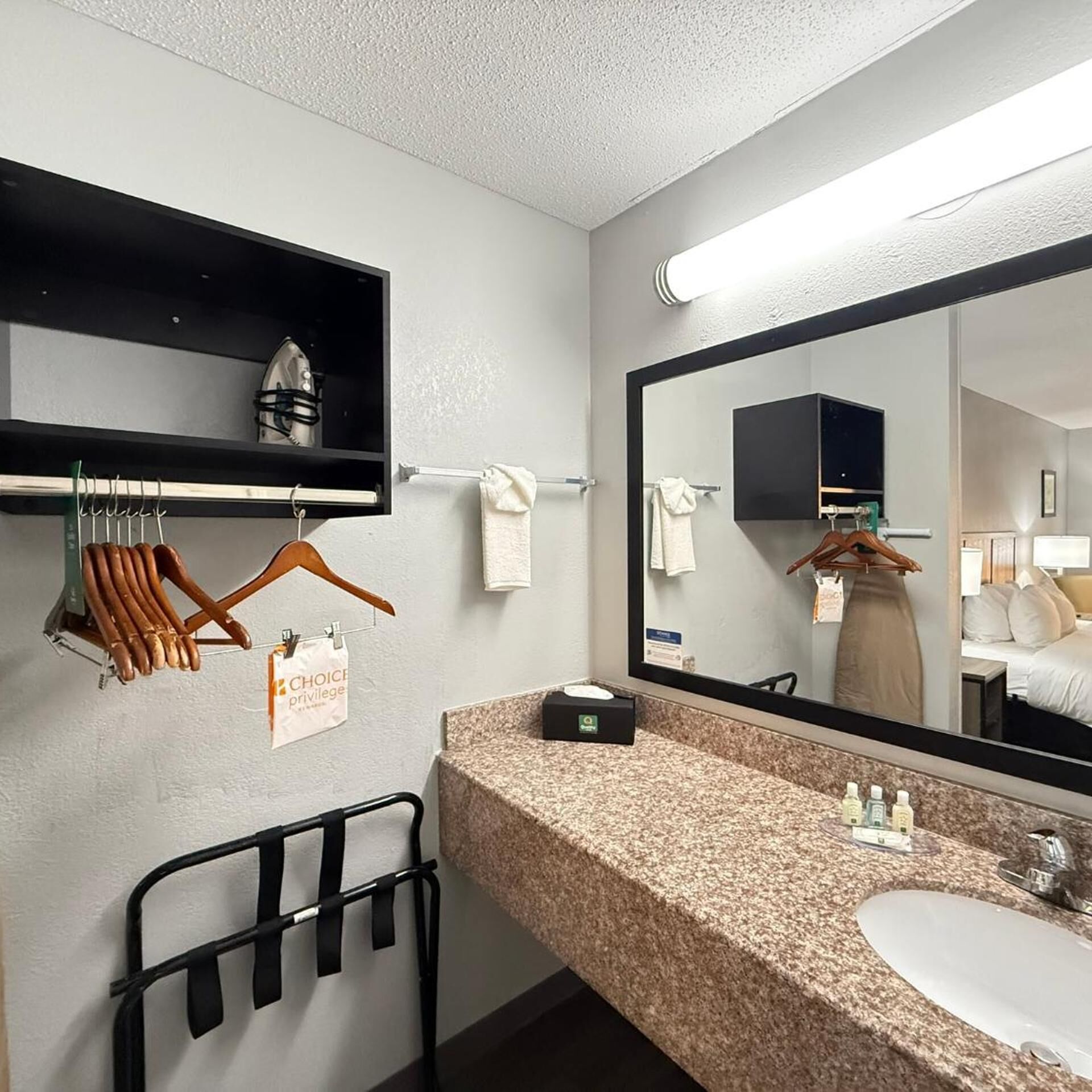 A hotel bathroom with a granite countertop, a sink, a mirror, a towel rack, and a clothing rack with a 'Choice Privilege' sign.