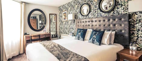 A hotel room with a large bed, a desk, a chair, a round mirror, and a wall with floral wallpaper.