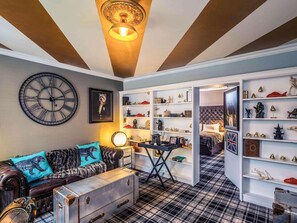 A room with a large clock, a sofa with patterned cushions, a table with a lamp, and a bookshelf with various items. - Nottingham City Centre Hotel by Sunday (Nottingham)