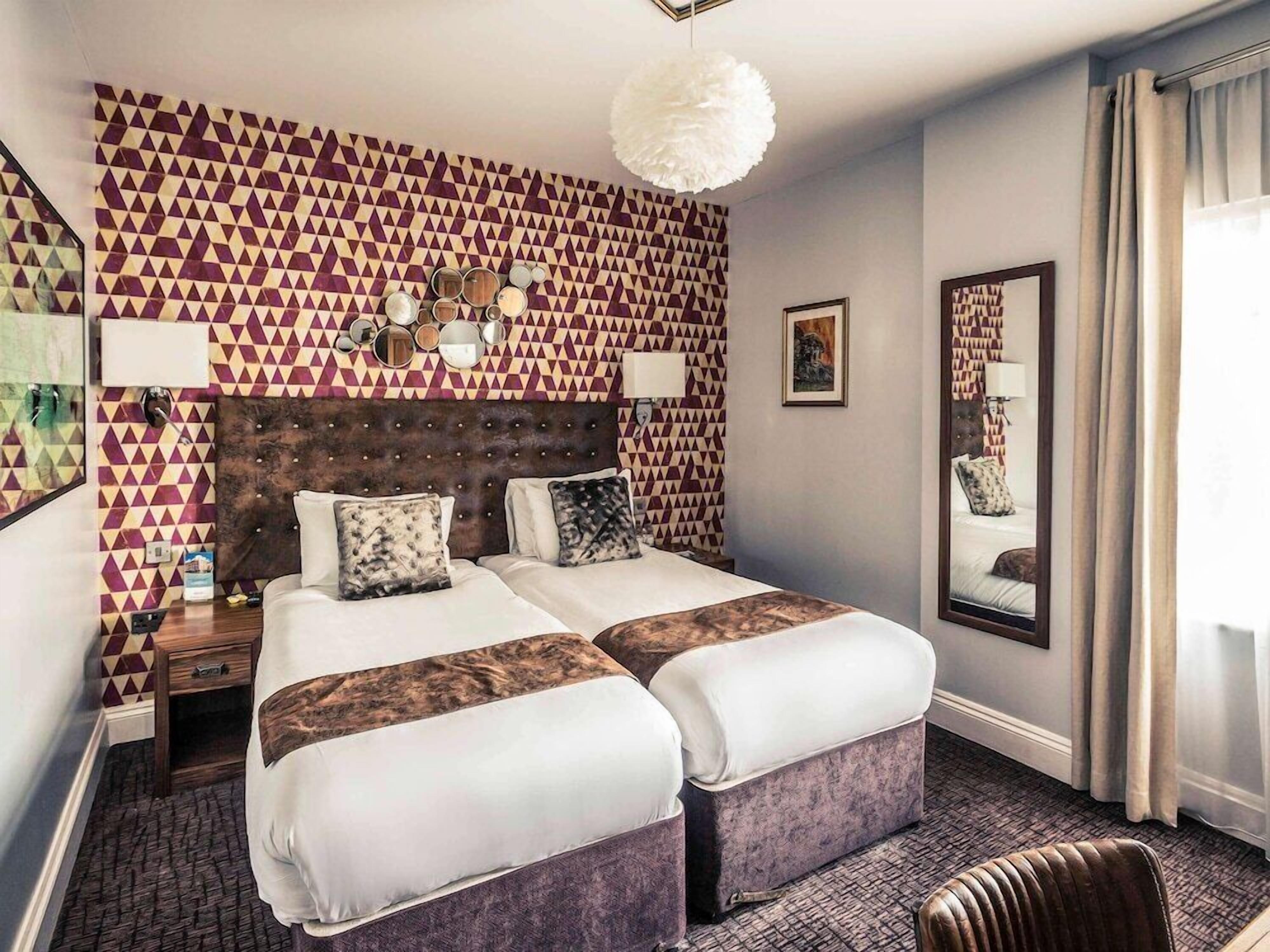 A hotel room with a geometric patterned wall, a tufted headboard, two beds with matching pillows, a large mirror, and a hanging light fixture.