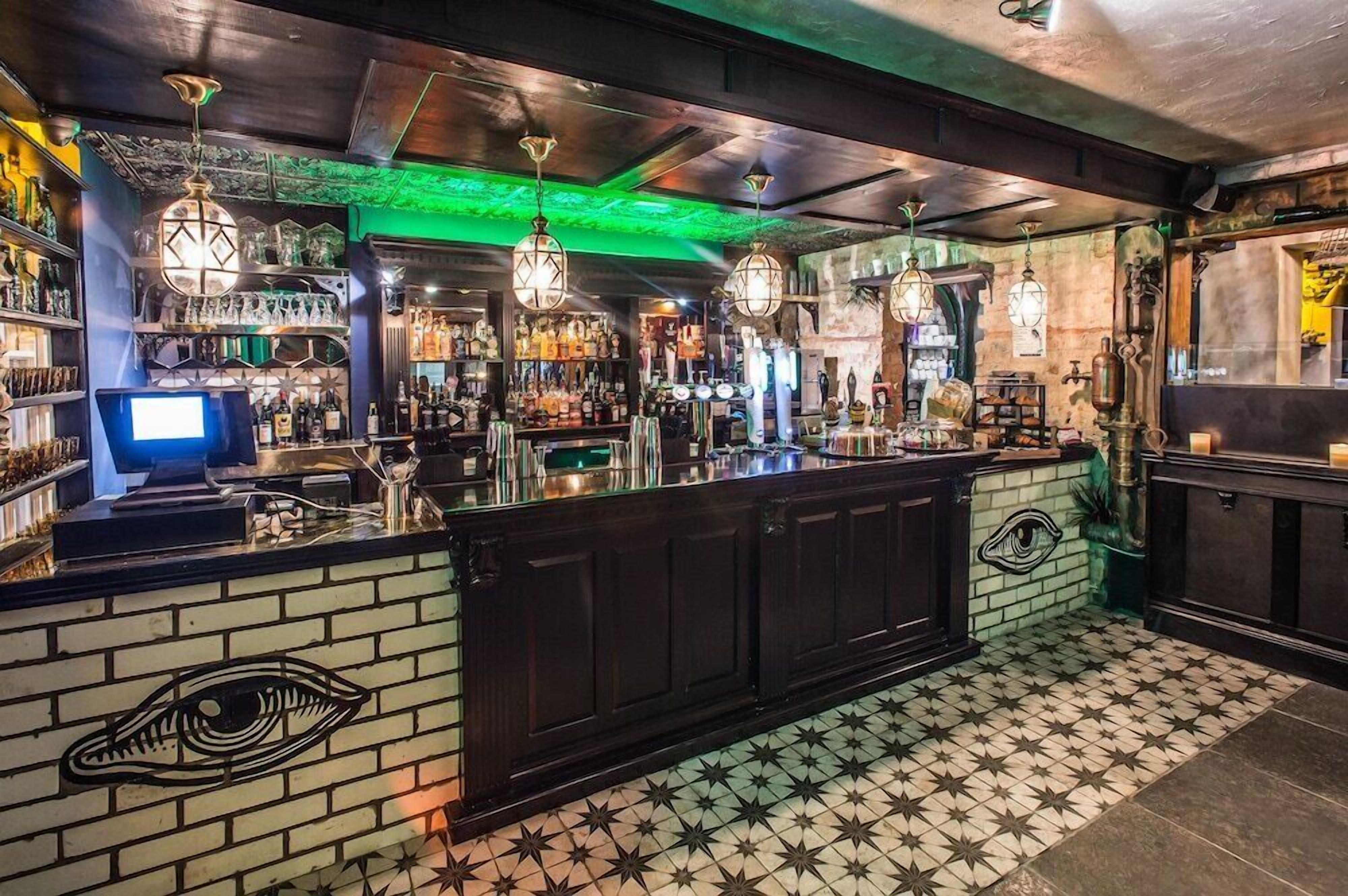 Una zona de bar con mostrador, estantes llenos de botellas, luces colgantes y un suelo estampado.
