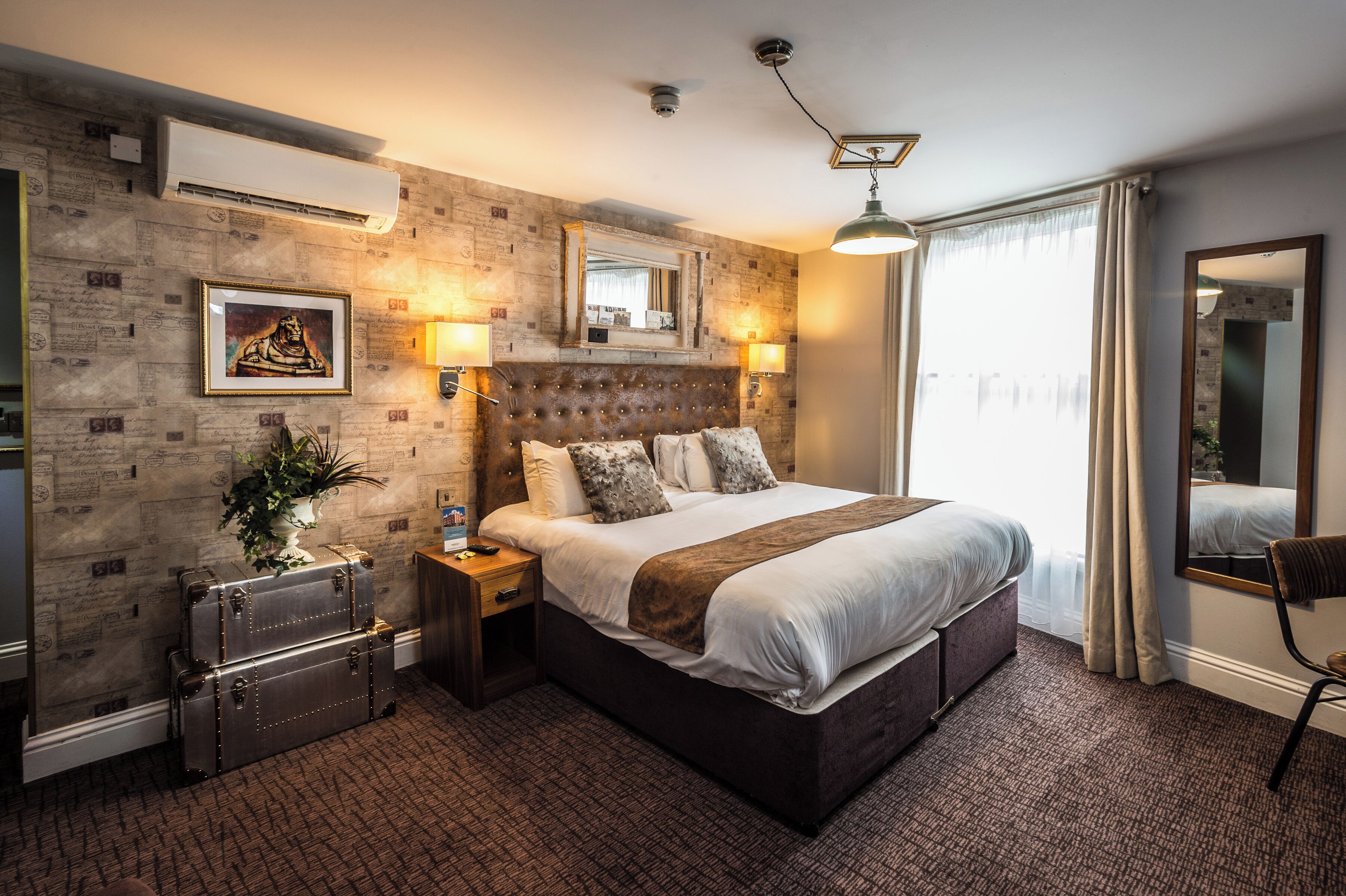 A hotel room with a large bed, a headboard, a nightstand, a suitcase, a plant, a mirror, and a window with curtains.
