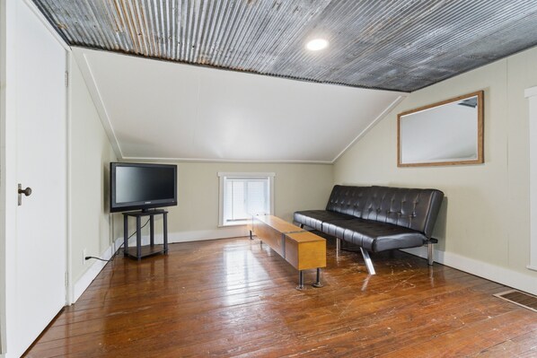 A room with a wooden floor, a black leather sofa, a TV on a stand, and a ceiling with exposed beams.