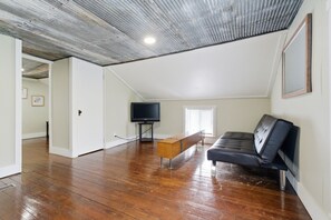 A spacious room with a wooden floor, a black sofa, a television, and a ceiling with exposed beams.