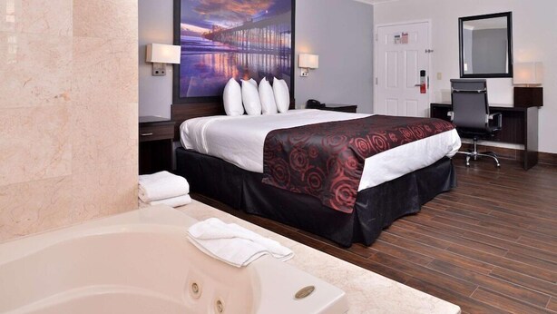 A hotel room with a large bathtub, a bed with a dark red patterned bedspread, a desk with a chair, and a large painting on the wall. - Emerald Inn on Sunset (Los Angeles)
