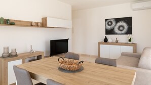 A modern living room with a wooden dining table, grey sofas, a flat-screen TV, and a wall-mounted artwork. - La Dimora Molara by Wonderful Italy (Olbia)