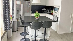 Ett modernt kök med en central köksö, barstolar, en hängande lampa och ett kylskåp.