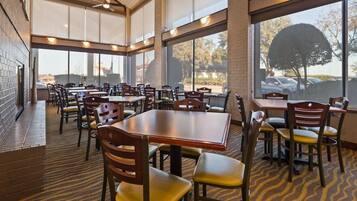 A spacious indoor dining area with multiple tables and chairs, large windows, and a brick wall.